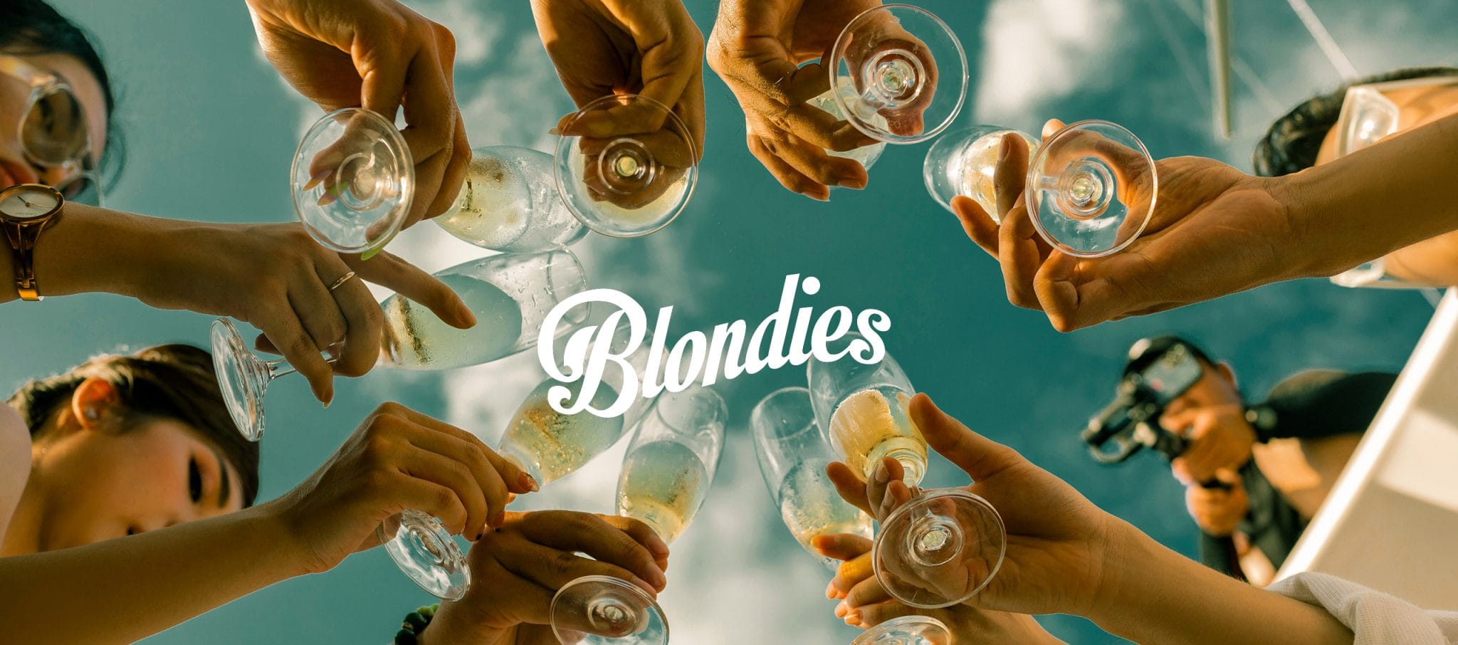 Blondies wordmark over a wide shot of friends toasting with coupe glasses against a blue sky