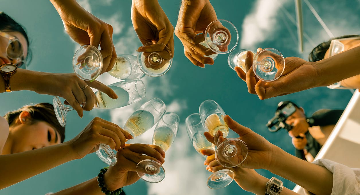 Friends clinking champagne glasses, photographed from below against a blue sky