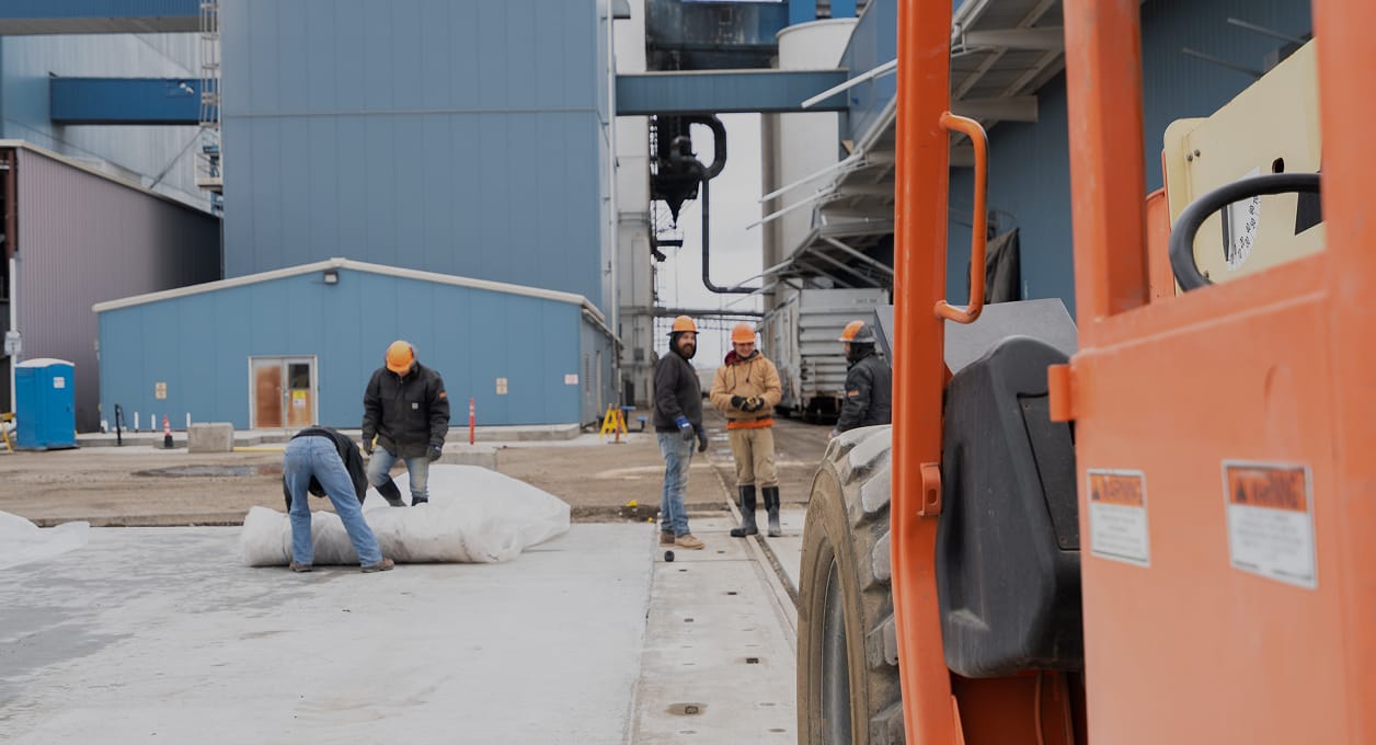 Construction crew in hard hats working beside a telehandler at a blue industrial plant